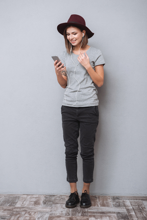Full length portrait of a smiling young woman in hat standing and listening music with earphones isolated on a gray backgroundの写真素材