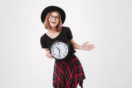 Portrait of a smiling cheerful woman in hat and eyeglasses holding wall clock isolated on a white backgroundの写真素材