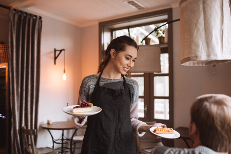 Picture of young lady confectioner with long hair standing in cafe while holding cake in hand. Look at client.の写真素材