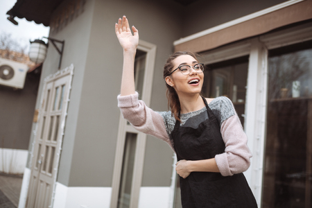 Image of pretty young woman confectioner with long hair standing near cafe while waving to friendsの写真素材