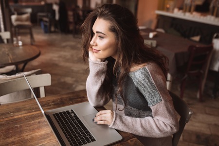Image of beautiful young woman with long hair sitting at the table in cafe while using a laptop. Look at laptop.の写真素材