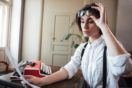 Side view of beauty authoress in white shirt holding her eyeglasses and sitting by the table with paper and typewriterの写真素材
