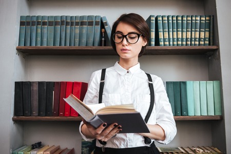 Authoress in glasses and white shirt reading book with bookshelf on backgroundの写真素材