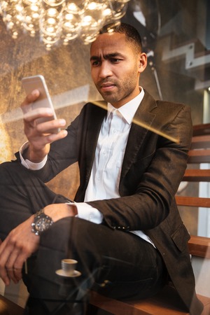 Vertical image of serious african man in suit using phone and sitting on stairs