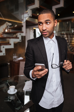 Vertical image of african man in suit with phone and glasses looking away in hotelの写真素材