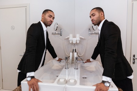 Side view of Young African man in suit looking at mirror in bathroom in hotel