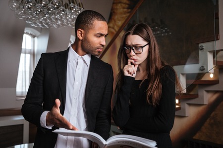 Serious African man in suit talking with woman in glasses and pointing at journal