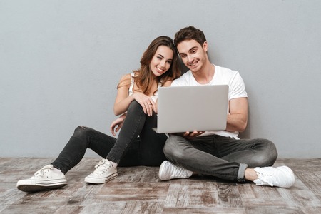Couple sitting on the floor with laptop computer in studio. Full length image. Isolated gray backgroundの写真素材