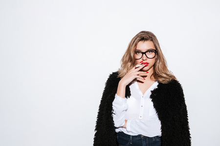 Portrait of a young stylish woman in fur coat and eyeglasses smoking cigarette and looking at camera on white backgroundの写真素材