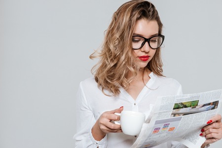Portrait of a smart young woman in eyeglasses reading newspaper and drinking cup of coffee isolated on a white backgroundの写真素材
