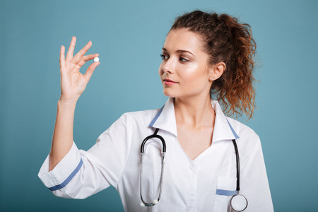 Young nurse wearing lab coat looking at pill in hand isolated over blue background.の写真素材