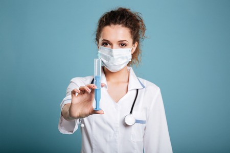 Woman in face surgical mask and white lab coat holding syringe isolated over blue background.の写真素材