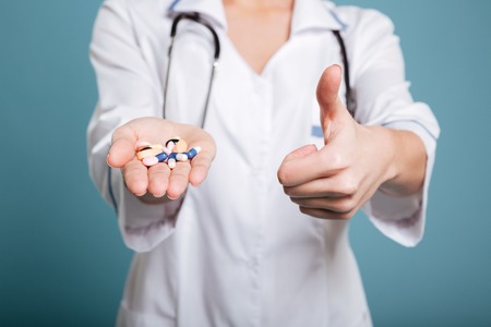 Woman nurse holding pills in hand and showing thumbs up isolated over blue background.の写真素材
