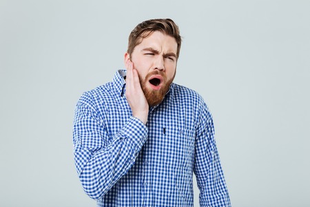 Depressed bearded young man shouting and having toothache over white backgroundの写真素材