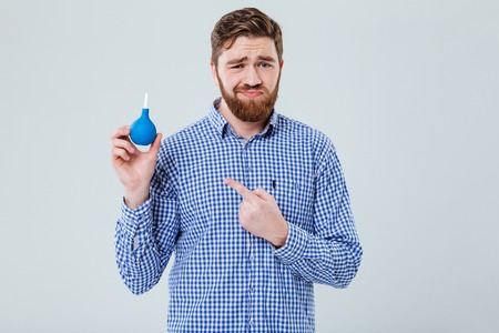 Unhappy frowning bearded young man holding enema and pointing on it over white backgroundの写真素材