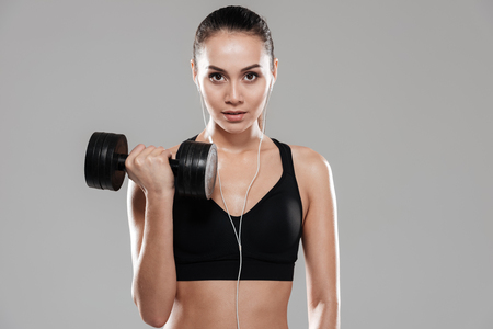 Front view of Sporting woman using dumbbell in studio and looking at camera. Isolated gray backgroundの写真素材