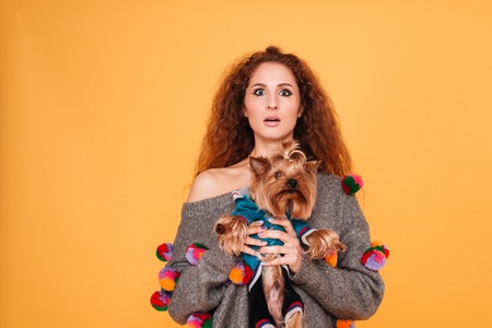 Young red head woman looking surprised while holding her dog isolated on orange backgroundの写真素材