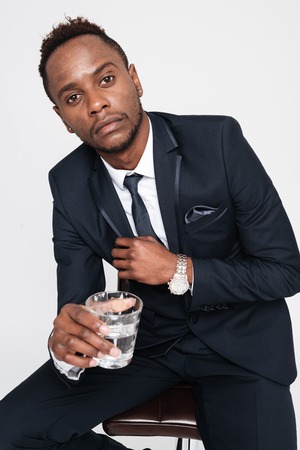 Image of african young businessman drinking water while sitting. Isolated over white background. Look at camera.の写真素材