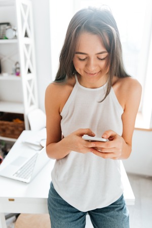 Image of pretty young woman standing at the home chatting by her phone near table. Look at phone.の写真素材