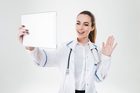Smiling pretty young woman doctor having video confetence with tablet over white background