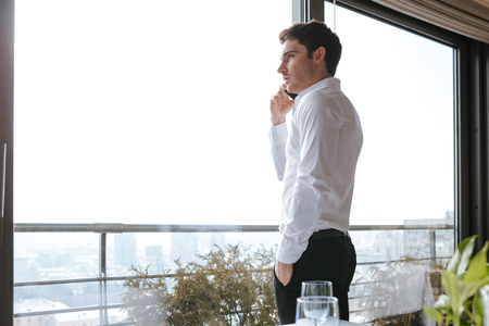 Side view picture of handsome serious young man dressed in white shirt standing in cafe indoors while talking by mobile phone. Looking aside.の写真素材