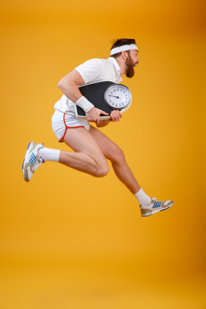 Image of screaming young sportsman jumping holding scales dressed in white t-shirt isolated over yellow background. Looking aside.の写真素材