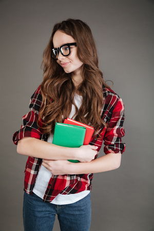 Photo of cheerful young woman wearing glasses posing isolated over grey background holding books in hands. Looking aside.の写真素材