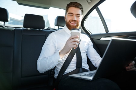 Smiling bearded business man sitting on back seat of a car with laptop and holding cup of coffee while looking at cameraの写真素材