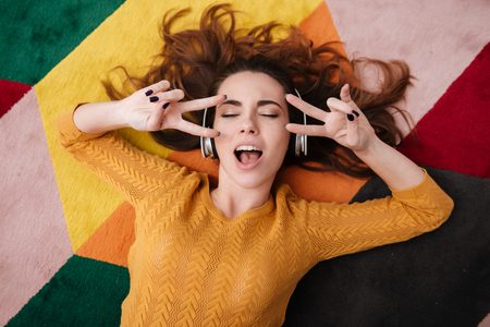 Top view portrait of a young pretty girl in headphones enjoying music while lying on a carpet and showing peace gesture at home with eyes closedの写真素材