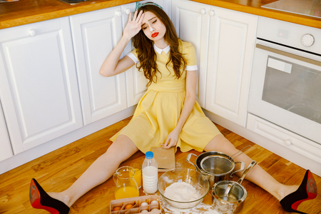 Image of sad tired young pin-up woman with red lipstick sitting on floor at kitchen and cooking. Looking aside.の写真素材