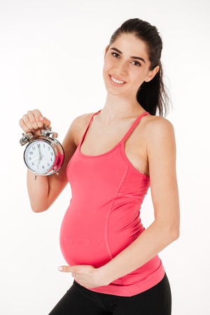 Smiling lovely pregnant woman holding alarm clock isolated on white background