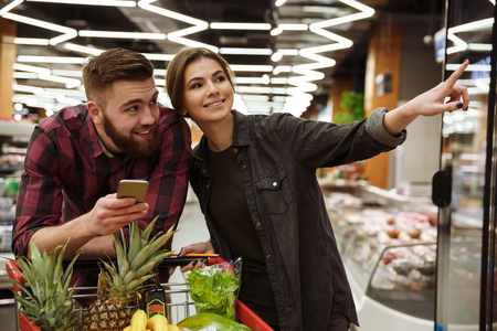 Image of young happy loving couple in supermarket with shopping trolley choosing products. Man holding mobile phone.の写真素材