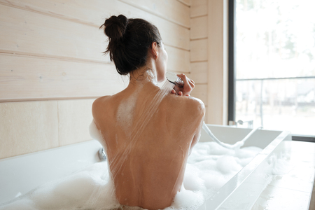 Back view of a naked young brunette woman washing in bathtub at homeの写真素材