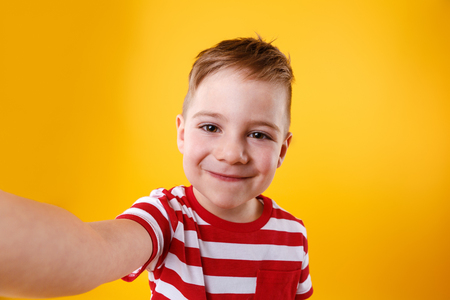 Portrait of a smiling little boy holding mobile phone and making selfie isolated over orange backgroundの写真素材
