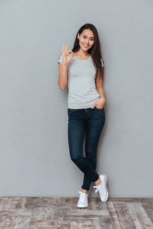 Vertical image of smiling woman showing ok sign and looking at camera while holding arm in pocket and posing in studio over gray backgroundの写真素材