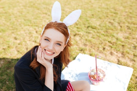 Portrait of a pretty young girl with long red hair wearing bunny ears and having easter picnic...