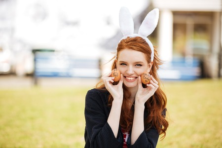 Portrait of a happy young woman with long ginger hair and bunny ears showing painted easter eggs outdoorsの写真素材