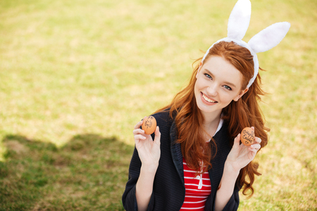 Portrait of a young smiling red head woman wearing bunny ears and showing two painted easter eggs...