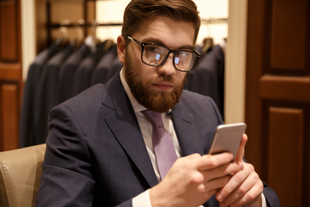 Photo of concentrated young bearded businessman sitting indoors and chatting by phone. Looking at camera.の写真素材