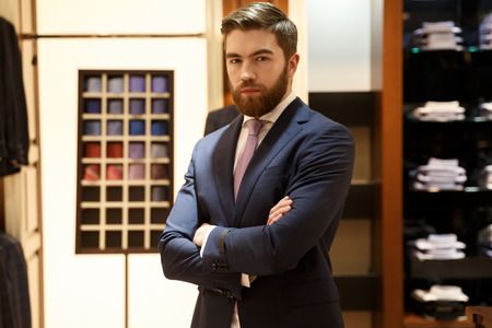 Calm bearded man in suit standing with crossed arms in a shop and looking at the cameraの写真素材