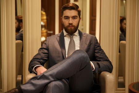 Serious man in suit sitting on armchair in a shop and looking at the cameraの写真素材