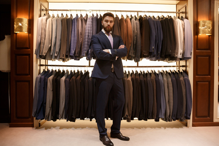 Full length portrait of bearded man in suit standing with crossed arms  near the rack with suits in a shop while looking at the cameraの写真素材