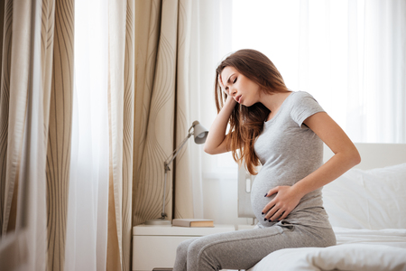 Sad pregnant young woman having headache and feeling sick on bed at homeの写真素材