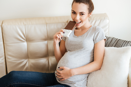 Smiling playful pregnant young woman eating chocolate bar on sofa at homeの写真素材