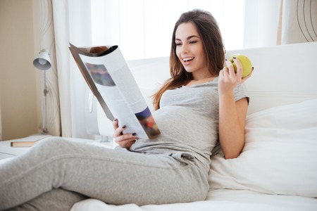 Cheerful pregnant young woman eating apple and reading magazine on bed at homeの写真素材