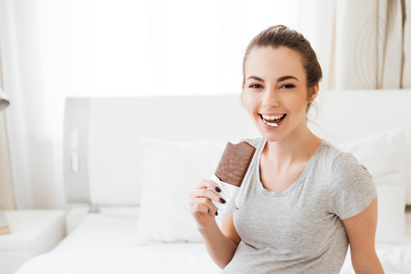 Cheerful pretty young woman eating chocolate bar and laughing on bed in bedroomの写真素材