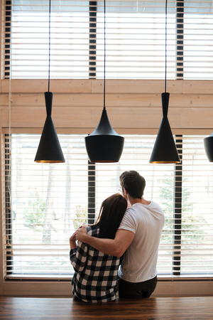 Back view of lovers young man and woman standing against window and huggingの写真素材