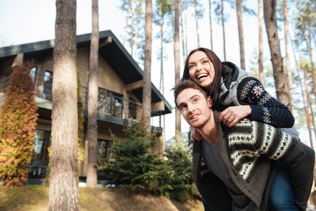 Cheerful woman riding on back of her smiling young man and looking away at forestの写真素材
