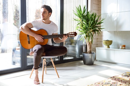 Young beatiful man with guitar in hands sitting on chair and looking away
