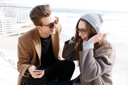 Cheerful young couple laughing and using smartphone together outdoors in autumnの写真素材